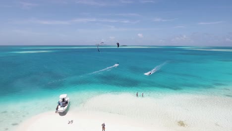 People-kitesurf-on-turquoise-clear-crystal-water-and-beach-camp-on-white-sand-beach,-drone-shot