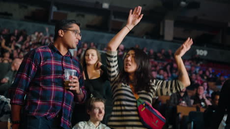 Young-Woman-Lady-in-Arena-Stands-Dancing-with-Arms-Raised-and-Hands-Up-During-Music-Show-Concert,-Boyfriend-Next-to-Her-and-Spectators-People-Around
