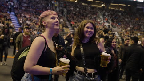 Two-Young-Ladies-Friends-Spectators-Standing-Dancing-Holding-Beer-Cups-Goblet-During-Music-Concert-Show,-Enjoying-The-Event,-Arena-Stands-and-Audience-Crowd-Around