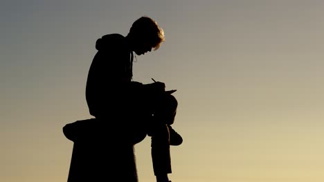 Silhouette-male-young-artist-painter-sitting-outside-looking-and-drawing-scene-in-front-of-him