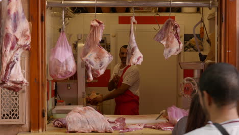 Carnicero-Preparando-Varias-Carnes-Frescas-Colgando-Fuera-Del-Mercado-De-Alimentos-De-Marrakech-Mientras-Los-Clientes-Pasan-Por-La-Ventana