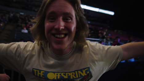 Joyful-Young-Guy-Entering-Arena-Before-Rock-Music-Concert-Show,-Smiling-Looking-At-Camera-With-Outstretched-Arms,-Spectators-Crowd-in-Stands-Around