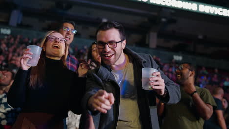 Joyful-Guy-Man-with-Girlfriend-Cheering-Singing-in-Arena-Stands-During-Music-Concert-Show,-Looking-at-Camera-With-Drink-Cup-in-Hand,-Spectators-Crowd-Around