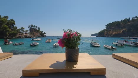 Dolly-shot-along-Flowerpots-on-beachfront-promenade,-Boats-moored-on-turquoise-water-bay,-Parga