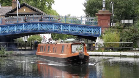 Narrow-boat,-which-is-also-a-houseboat,-sailing-under-blue-bridge-at-Little-Venice,-London,-England