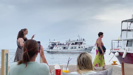 June-2022-Unidentified-Tourists-Enjoying-a-Drink-at-a-Greek-Island-Harbour-Videoing-The-View-With-People-Walking-Past-And-Boats-In-The-Harbour