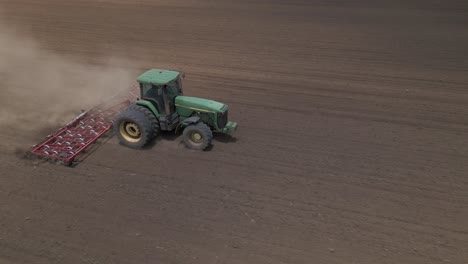 Green-tractor-pulling-harrow-in-field-kicks-up-clouds-of-dust,-aerial