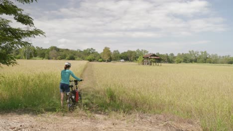 A-stationary-footage-of-a-lone-female-cyclist-walking-her-bike-going-into-a-rice-farm-or-wheat-field