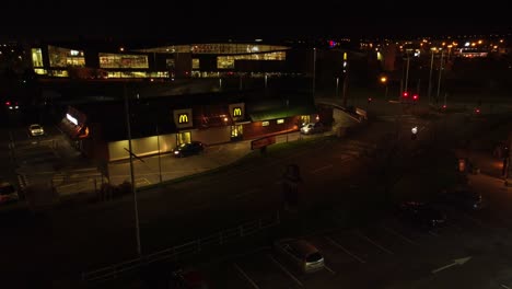 Flying-across-McDonalds-fast-food-drive-through-illuminated-at-night-alongside-UK-town-highway-aerial-descending-view