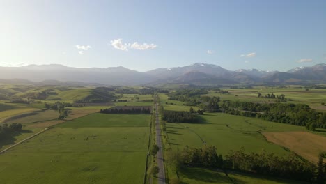 Circling-above-a-straight-road-that-leads-towards-a-mountain-range-during-beautiful-golden-hour-light-in-the-countryside-of-New-Zealand
