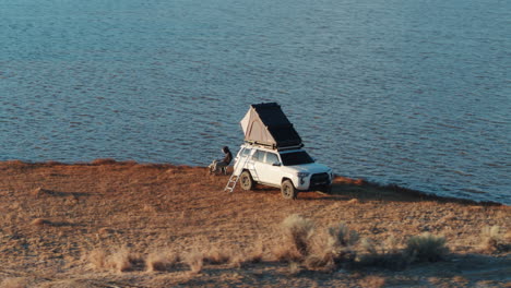 White-car-with-roofnest-tent-and-man-sitting-next-to-lake,-aerial