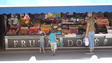Dad-with-kids-at-spanish-fruit-market-stall-by-sidewalk-on-street-on-a-sunny-day