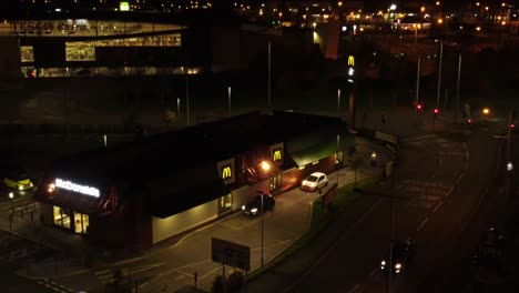 Cars-queue-outside-McDonalds-fast-food-drive-through-at-night-in-Northern-UK-town-aerial-zoom-out-view