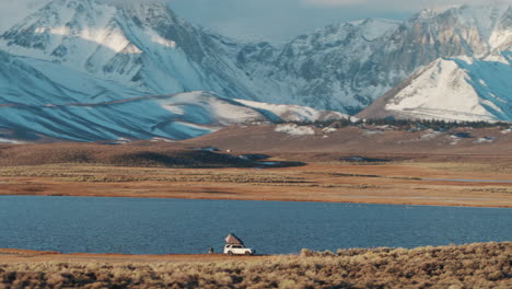 Remote-wild-camping-on-shore-of-valley-lake-with-snowy-mountains,-aerial