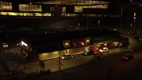 Cars-queue-outside-McDonalds-fast-food-drive-through-illuminated-at-night-in-Northern-UK-town-aerial-orbiting-view