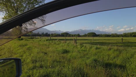 Farmland-and-snow-capped-mountains-viewed-through-open-car-window-during-sunset