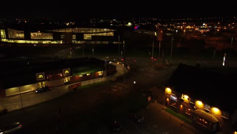 Flying-over-McDonalds-fast-food-drive-through-illuminated-at-night-alongside-UK-town-highway-aerial-view-descending-left
