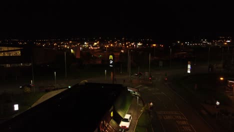 Flying-over-McDonalds-fast-food-drive-through-illuminated-at-night-alongside-UK-town-highway-aerial-view