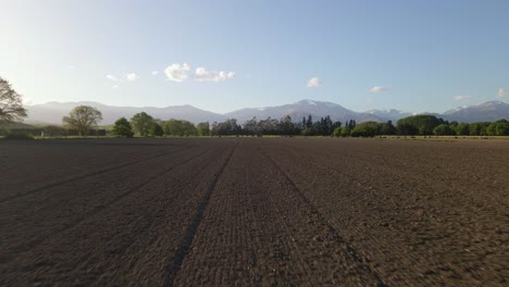 Low,-fast-drone-flight-over-freshly-ploughed-farm-land-during-sunset,-mountain-range-in-background