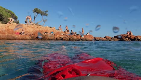 Slow-motion-personal-perspective-of-man-wearing-red-swimwear-with-legs-and-feet-relaxing-while-floating-on-calm-sea-water-with-Palombaggia-rocks-and-people-in-background
