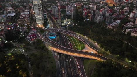 Aerial-view-in-front-of-traffic-at-the-Petroleos-Fountain,-roundabout-and-junction,-dusk-in-Mexico-city---ascending,-drone-shot