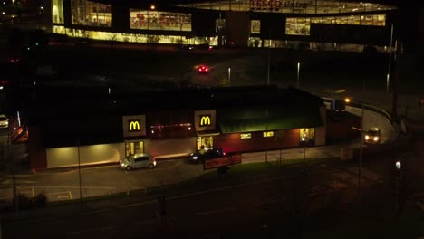 Vehicles-queue-outside-McDonalds-fast-food-drive-through-illuminated-at-night-in-Northern-UK-town-aerial-orbiting-view