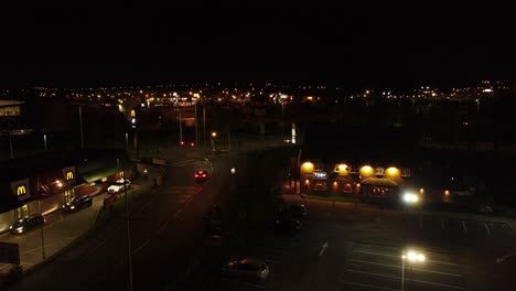 Flying-over-McDonalds-fast-food-drive-through-illuminated-at-night-alongside-UK-town-highway-aerial-view-rising-left