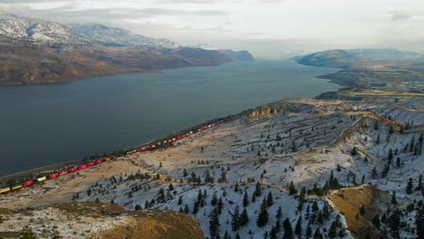 An-extraordinary-capture-of-a-colorful-train-passing-by-Kamloops-Lake-in-the-winter,-a-hilly-desert-terrain-with-spruce-trees-in-the-foreground