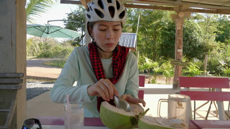 Young-Asian-woman-wearing-cycling-helmet-enjoying-eating-tender-coconut-pulp-using-a-spoon-in-a-village-in-Northern-Thailand