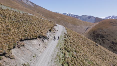 Aerial-view-tracking-three-hikers-up-a-winding-dirt-road-in-the-alps-of-New-Zealand-on-a-sunny-spring-day