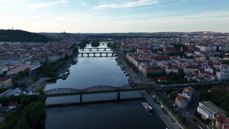 A-drone-view-of-Prague-during-sunset---flying-over-the-Vltava-river-towards-Prague-Castle,-train-crossing-the-bridge