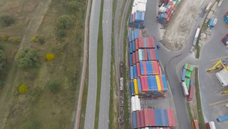 Aerial-view-train-pulls-wagons-with-cargo-parallel-to-car-road-and-cargo-terminal-in-Klaipeda-city