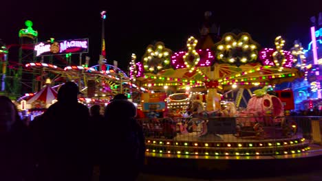 Pan-shot-of-locals-walking-around-a-merry-go-round-in-a-amusement-park-in-London,-UK-at-night-time