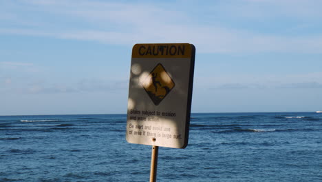 Erosion-warning-sign-on-Waikiki-beachfront,-Ocean-Waves-in-Background,-Handheld