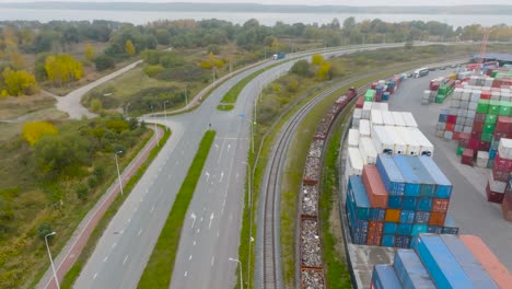 Aerial-view:-a-train-pulls-cargo-wagons-parallel-to-a-car-road-and-a-cargo-terminal-in-Klaipeda-city