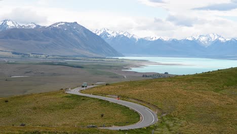 Farm-truck-driving-down-winding-road-with-stunning-alpine-mountain-range-in-background-and-turquoise-lake-Tekapo
