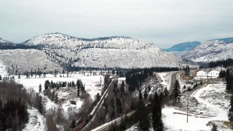 Frosty-British-Columbia:-Aerial-Tracking-Shot-of-Autos-on-Yellowhead-Highway-5-and-Freight-Train-on-Rail-Through-Icy-North-Thompson-River-Valley-near-Kamloops,-Snow-covered-Landscape-Wide-View