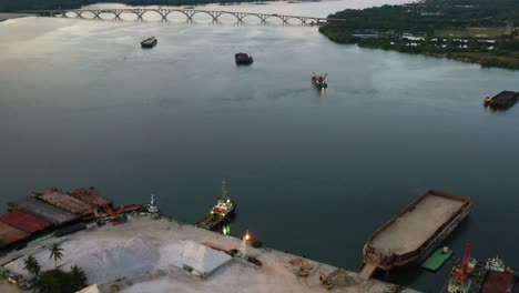 Aerial-flyover-Lee-Trading-Company-at-lumut-port-industrial-park-with-sand-and-gravel-piles-at-the-site,-tilt-up-reveals-permaisuri-bainun-bridge-across-manjung-river,-Kampung-Acheh,-Malaysia