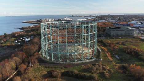 Rotating-aerial-shot-of-a-disused,-out-of-commission-Gasometer-by-the-sea-on-a-sunny-day