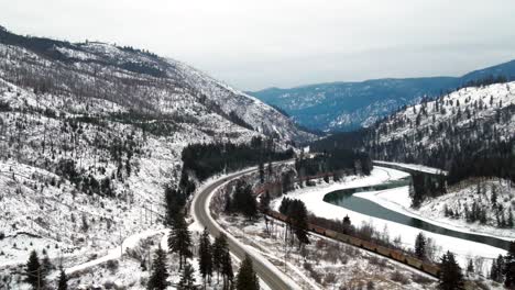 Aerial-Tracking-Shot-of-Autos-on-Yellowhead-Highway-5-and-Cargo-Train-on-Rail-Through-Partially-Iced-North-Thompson-River-Valley-near-Kamloops,-Snow-covered-Landscape-Wide-View