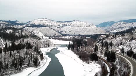Wide-Aerial-View-of-Vehicles-and-Transport-Train-on-Yellowhead-Highway-5-and-Railway-Through-Partially-Frozen-North-Thompson-River-Valley-near-Kamloops,-Snowy-Landscape-Panoramic-View