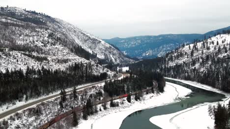 Frozen-North-Thompson-River-Valley-near-Kamloops:-Panoramic-View-of-Mountainous-Snow-covered-Landscape-with-Cars-on-Yellowhead-Highway-5-and-Freight-Train-on-Railway-in-Wide-Aerial-Shot-in-BC