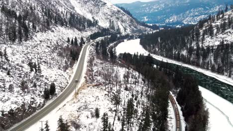 A-Medium-Aerial-Shot-of-Cars-and-Freight-Train-on-Yellowhead-Highway-5-and-Railway-Through-the-Frozen-North-Thompson-River-Valley-near-Kamloops,-Magnificent-Mountainous-Snow-covered-Landscape