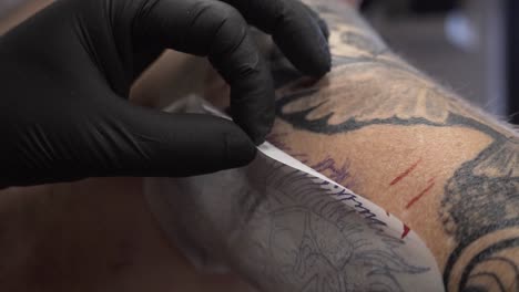 Artista-Del-Tatuaje-Escondió-Las-Manos-Con-Guantes-Negros-Tirando-Del-Papel-De-Preparación-Del-Tatuaje