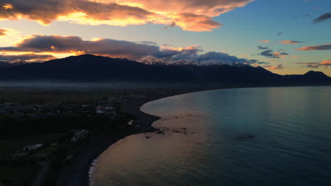Drone-shot-of-sea-with-lapping-waves-and-white-snow-capped-mountain-background-houses-orange-dusk-dawn-sky-new-Zealand