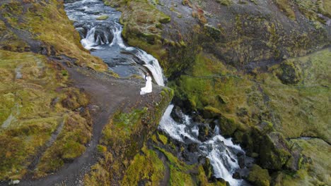 Newlywed-Caucasian-Couple-Hugging-And-Standing-On-Cliff-Next-To-Gorgeous-Waterfall-In-Iceland-Landscape