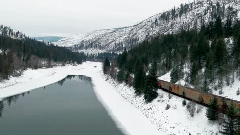 Close-Up-of-Freight-Train-Traveling-Through-a-Snowy-Landscape-in-British-Columbia:-Aerial-Shot-Follows-Journey-Along-the-Partially-Frozen-North-Thompson-River-and-Yellowhead-Highway-5-Near-Kamloops