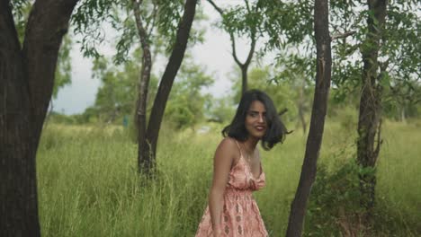 An-attractive-Asian-female-turning-around-looking-back-smiling-with-joy-and-excitement-while-wandering-through-a-grass-field-surrounded-by-trees,-India