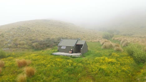 Person-sitting-on-small-deck-in-front-of-hiker's-hut-within-a-flower-field-in-the-New-Zealand-wilderness