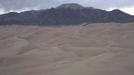 People-walk-across-desolate-sand-dune-field-with-mountains-in-back-ground